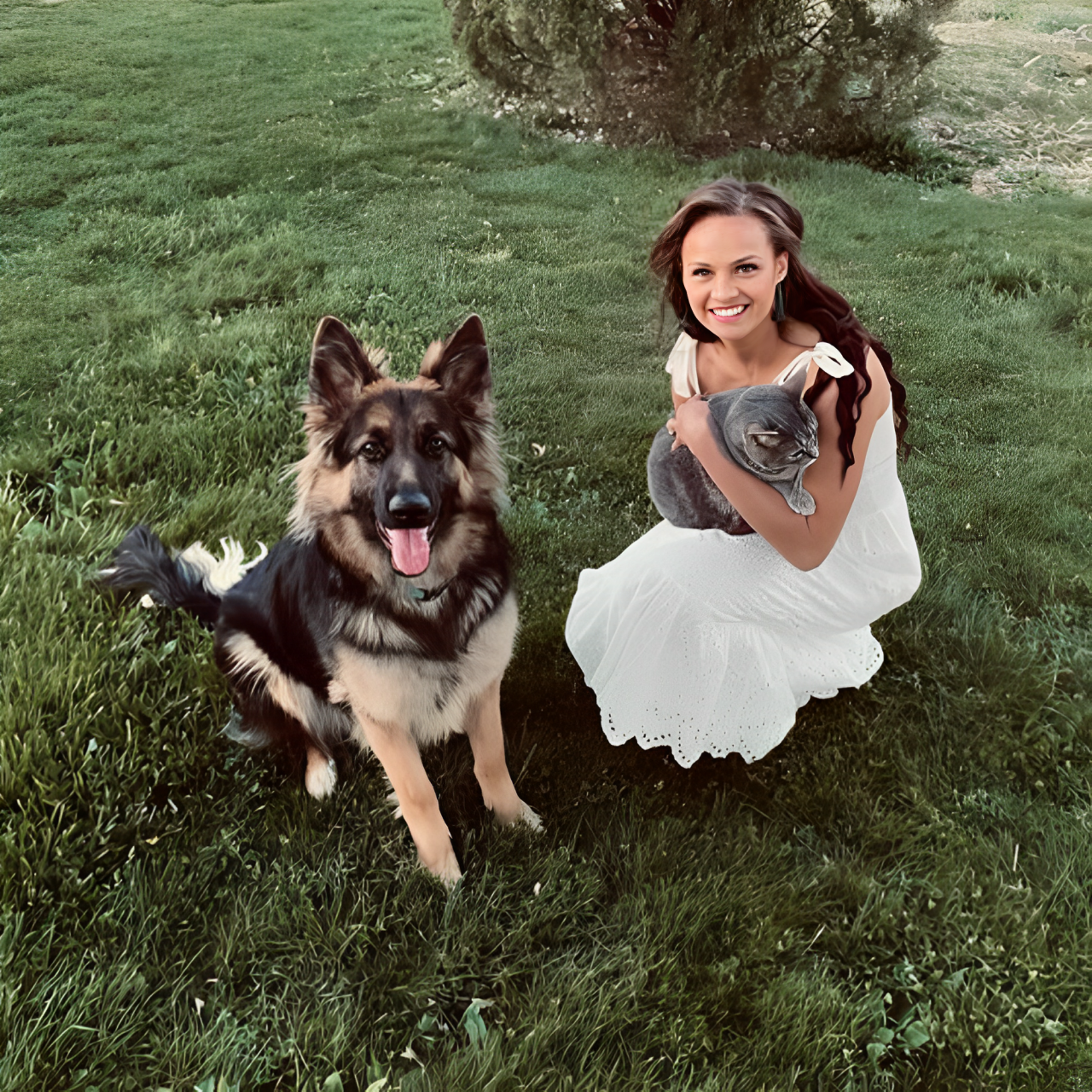 A woman sitting on the grass with her dog and cat, and wearing a white romper pocket dress.